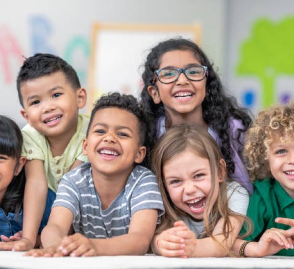 istockphoto-1166892023-612x612 Portrait of a happy multi-ethnic group of kindergarten age students. The cute children are laying in a pile on the ground in a modern classroom. The kids are laughing and smiling.